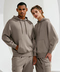 Two people, one male and one female, are standing side by side wearing matching beige TR602 hoodies and pants from TriDri. They both have their hands in their pockets and are looking directly at the camera.