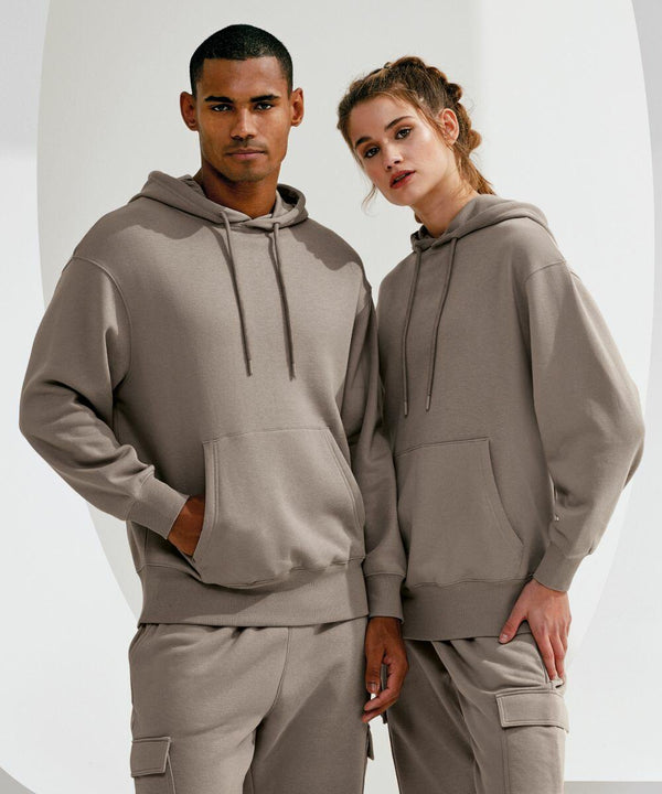 Two people, one male and one female, are standing side by side wearing matching beige TR602 hoodies and pants from TriDri. They both have their hands in their pockets and are looking directly at the camera.