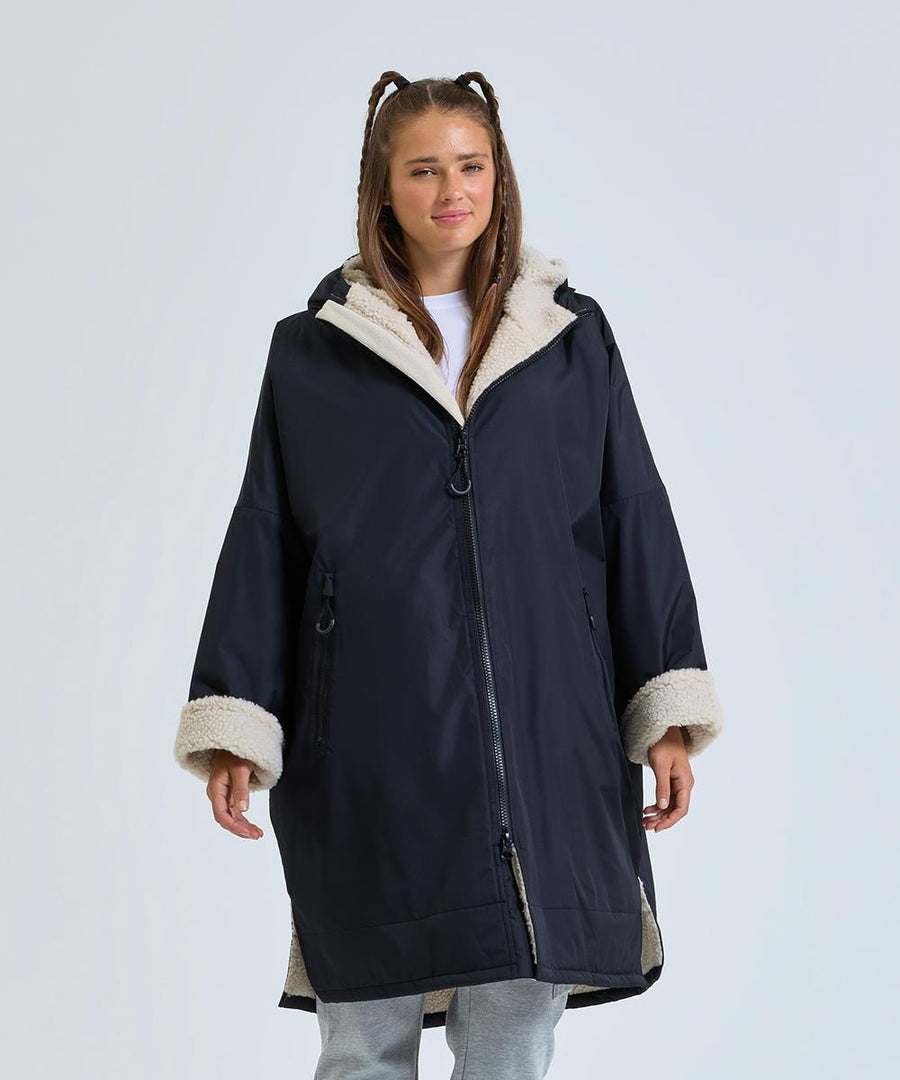 A person wears a TriDri All-seasons waterproof changing robe by TriDri, featuring a long, dark design with light fleece lining on the hood, cuffs, and interior. The robe is unzipped at the bottom and has front pockets, set against a plain light background.