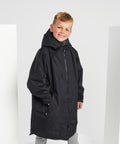 A blond-haired boy smiles in front of a white background with props, wearing an oversized black TriDri Kids TriDri All-seasons waterproof changing robe.