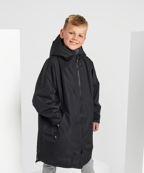 A blond-haired boy smiles in front of a white background with props, wearing an oversized black TriDri Kids TriDri All-seasons waterproof changing robe.