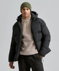 A person wearing the 2786 Recycled TrailPeak padded jacket, a beige knitted jumper, olive green beanie, and dark trousers stands against a plain light grey background.
