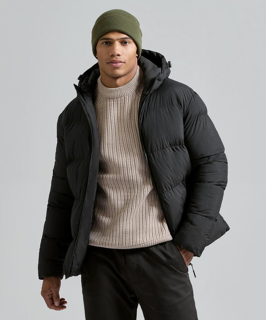 A person wearing the 2786 Recycled TrailPeak padded jacket, a beige knitted jumper, olive green beanie, and dark trousers stands against a plain light grey background.