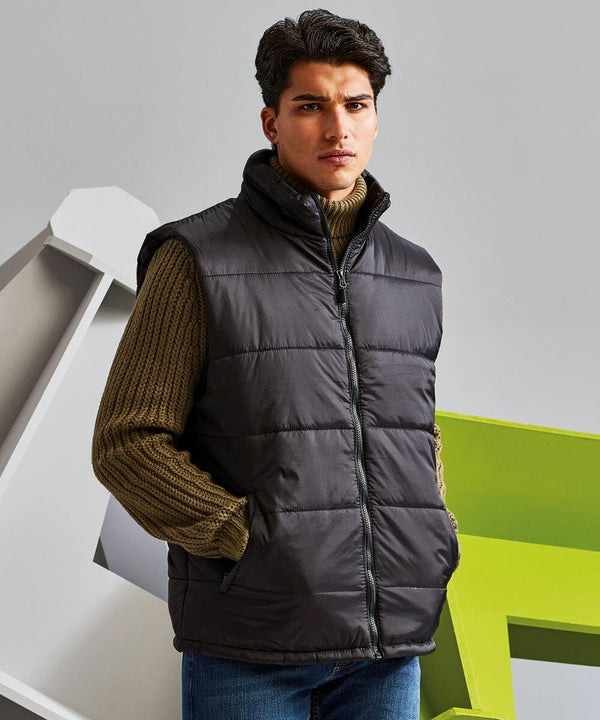 A man with dark hair is wearing a green knitted sweater and a black 2786 TS015 puffer vest. He is standing against a geometric background with white and green shapes.