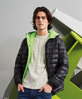 A person with shoulder-length hair wearing a black 2786 TS016 puffer jacket with a green lining over a white knitted sweater, standing in front of an abstract geometric structure.