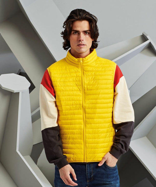 A person with medium-length hair wears a yellow quilted jacket from 2786 over a colorful long-sleeve shirt, standing against a backdrop with geometric shapes.
