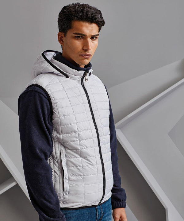 A person with dark hair is standing, wearing a white quilted vest from 2786 over a dark long-sleeve top, in front of a geometric background.