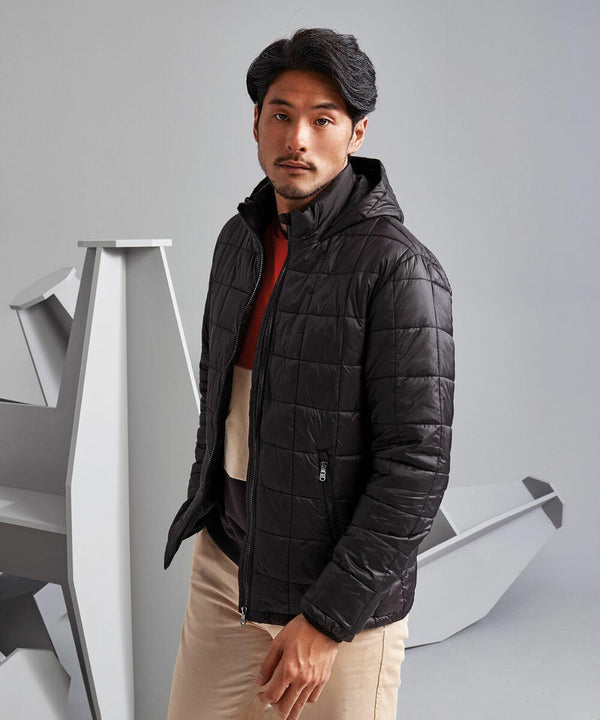 A man with a beard and mustache is standing in front of abstract geometric shapes. He is wearing the TS025 by 2786, a black quilted jacket, over a layered outfit consisting of a top and light-colored pants.