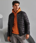 A man is standing against a neutral background, wearing an orange hoodie under a black 2786 TS030 puffer jacket and grey pants. He looks to his left with a serious expression.