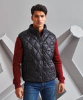 A man stands against a geometric background, wearing a black quilted vest from 2786 over a red long-sleeve shirt and blue jeans.