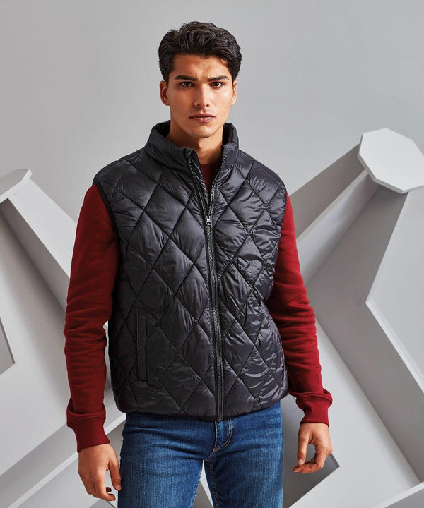 A man stands against a geometric background, wearing a black quilted vest from 2786 over a red long-sleeve shirt and blue jeans.