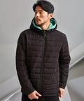 A man wearing a black 2786 TS043 with a hood and a green hooded sweatshirt underneath stands indoors against a grey background.