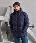 A man in a dark blue TS045 puffer jacket by 2786 with his hands in the pockets stands against a backdrop with geometric shapes.