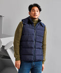 A man wearing a green knit sweater, blue puffer vest (2786 TS046), and blue jeans stands in front of a minimalist gray background.