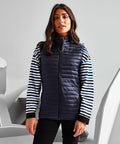 A woman stands wearing a navy 2786 TS19F quilted vest over a striped long-sleeve shirt, with a neutral expression. The background is minimalistic and monochromatic.