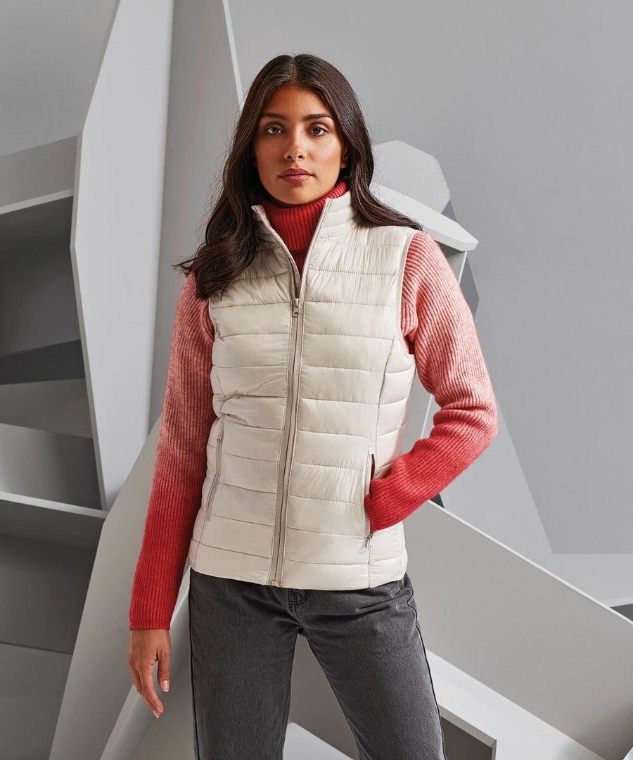 A woman with long dark hair wears a white 2786 TS31F quilted vest over a pink sweater and grey jeans, standing against a geometric grey backdrop.