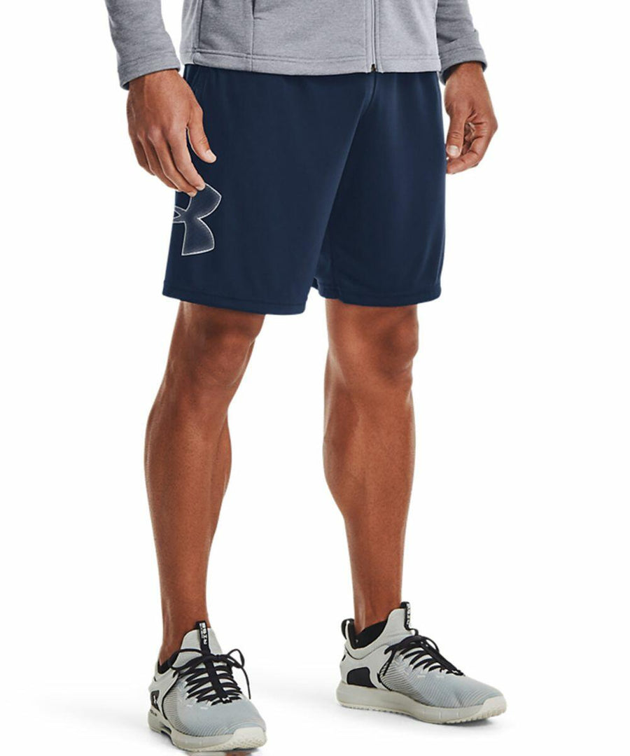 A person is shown from the waist down wearing a gray jacket, navy blue shorts with an Under Armour logo on the left thigh, and gray UA017 athletic shoes with white soles.