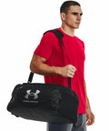A man in a red shirt and gray pants carries a black Under Armour UA032 over his shoulder.