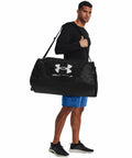 A man stands holding a large black Under Armour UA033 duffel bag over his shoulder. He is wearing a long-sleeved black shirt, blue shorts, and black sneakers.