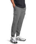 A person wearing Under Armour UA Vibe cargo trousers in grey with side flap pockets and elasticated cuffs, paired with a black shirt and white trainers, stands with hands in their pockets.