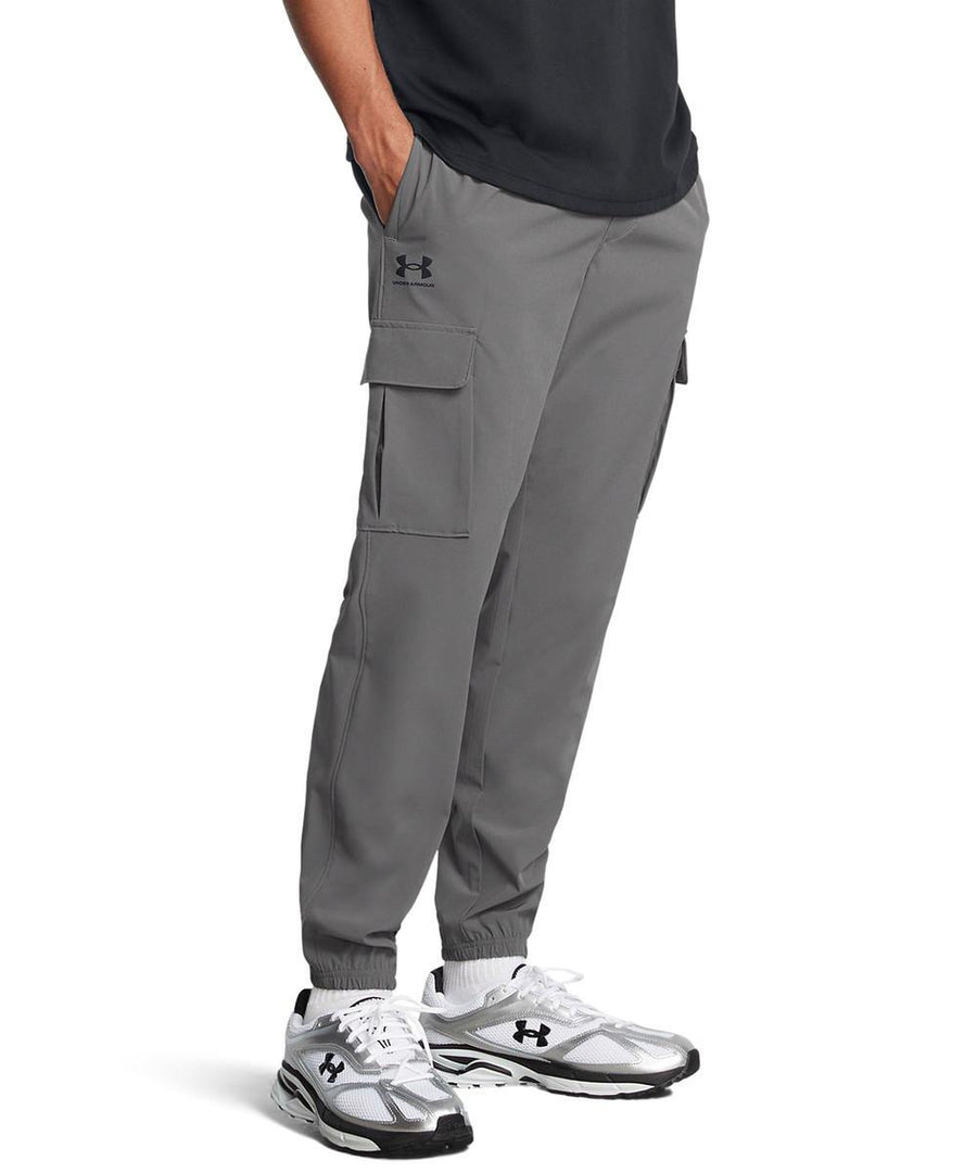 A person wearing Under Armour UA Vibe cargo trousers in grey with side flap pockets and elasticated cuffs, paired with a black shirt and white trainers, stands with hands in their pockets.