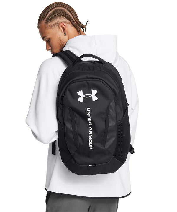 A person in a white hoodie and grey trousers is seen from behind carrying a black Under Armour Hustle 6.0 rucksack with the brand’s name and logo in white on the back.