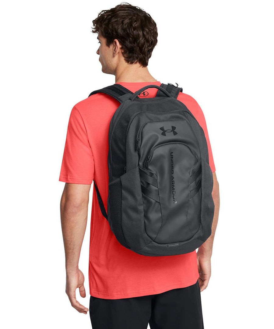 A person in a bright coral short-sleeve shirt and black shorts is seen from the back, carrying an Under Armour Hustle 6.0 Pro rucksack with a visible logo and front zip pocket.