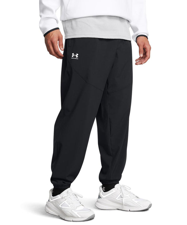 A person wearing Under Armour Vibe woven joggers in black, paired with a white long-sleeve top and white trainers, stands against a plain white background. Only the lower half of their body is visible.