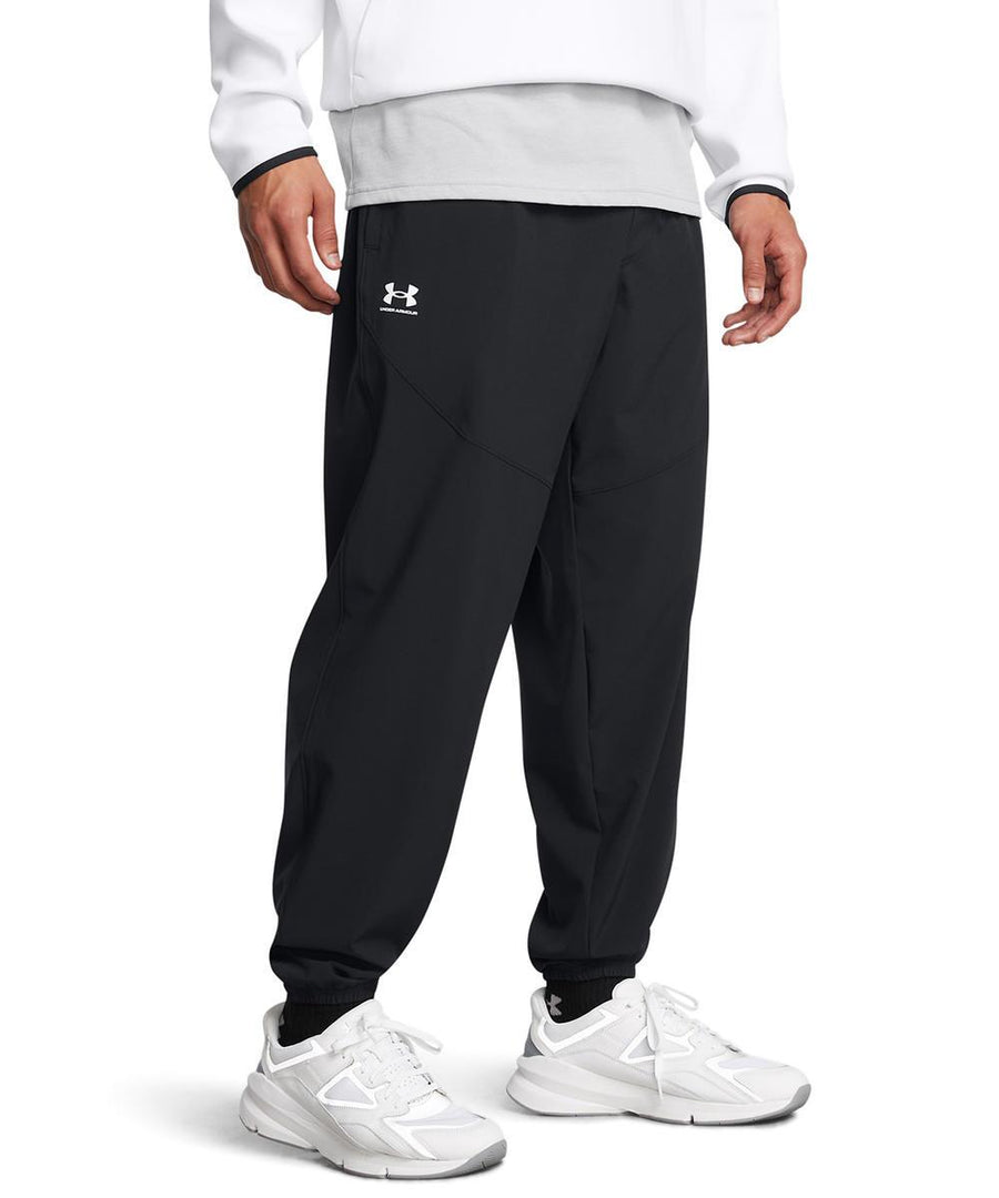 A person wearing Under Armour Vibe woven joggers in black, paired with a white long-sleeve top and white trainers, stands against a plain white background. Only the lower half of their body is visible.