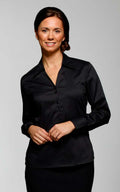 A woman with dark hair pulled back stands against a plain background. She is wearing a black, long-sleeve button-up Vortex freya blouse and is smiling at the camera.