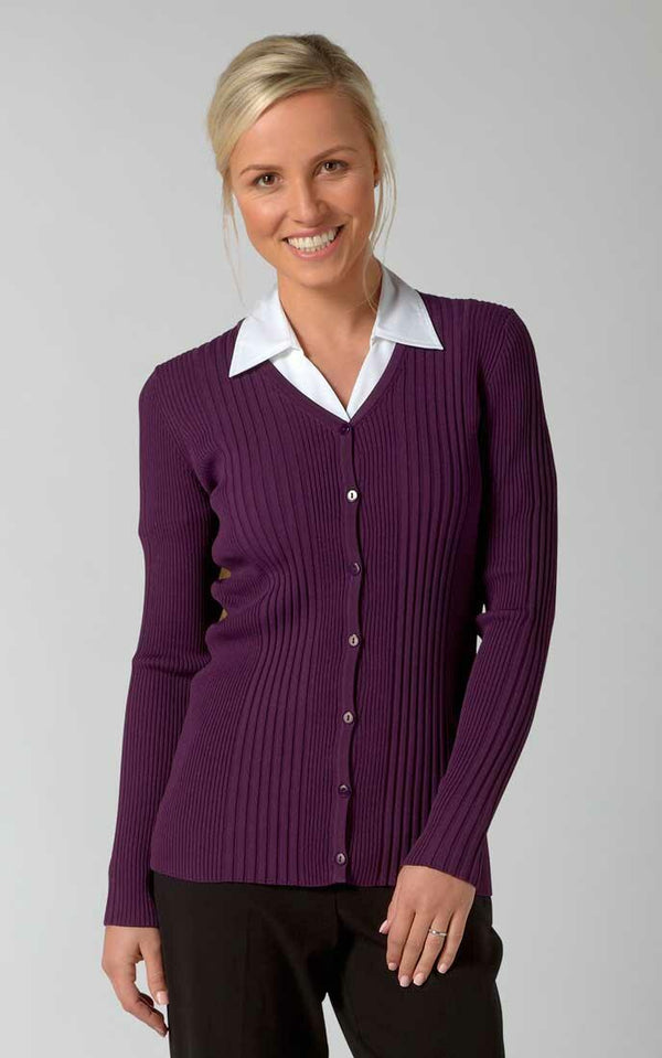 A person with blonde hair, wearing a white collared shirt under a purple, ribbed, button-up kristin by Vortex and black pants, smiles while standing against a plain, light gray background.