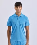 A young man faces forwards against a plain background, wearing the Wombat Men's Terry polo in light blue paired with matching blue trousers.