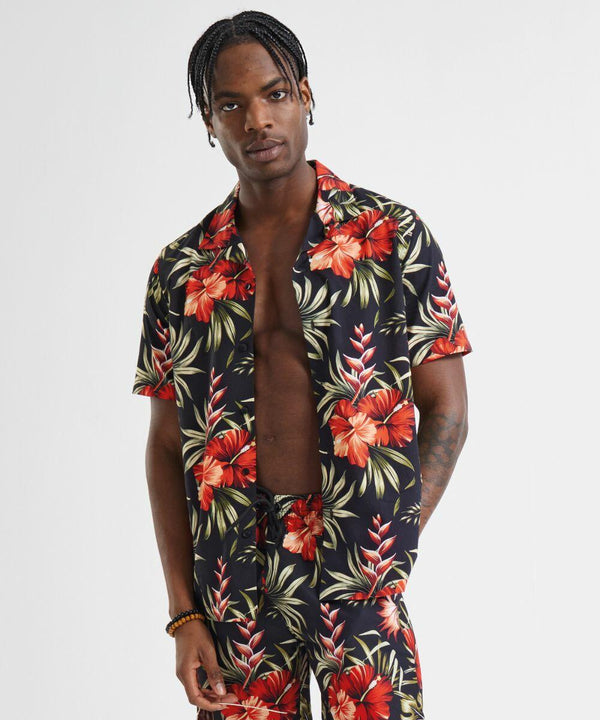 A person with braided hair wears a black, floral-patterned Wombat WB500 short-sleeve shirt and matching pants. The shirt is open, revealing their bare chest. They stand against a plain white background.