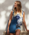 A person with long hair wearing a tie-dye sleeveless shirt and denim shorts leans against a white wall while holding a blue Westford Mill WM101 tote bag.