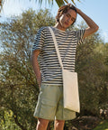 A person with shoulder-length hair wearing a striped t-shirt, beige shorts, and a white Westford Mill WM107 stands outdoors, with trees and a blue sky in the background.