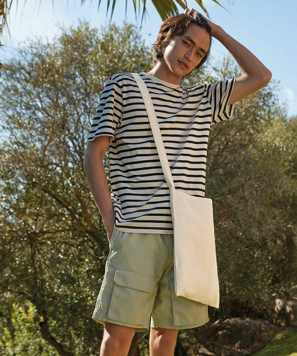 A person with shoulder-length hair wearing a striped t-shirt, beige shorts, and a white Westford Mill WM107 stands outdoors, with trees and a blue sky in the background.