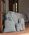 Four gray Westford Mill WM115 drawstring bags of varying sizes are placed on a tiled floor near a partially open wooden door. Behind, a bed with a striped bedspread is visible.