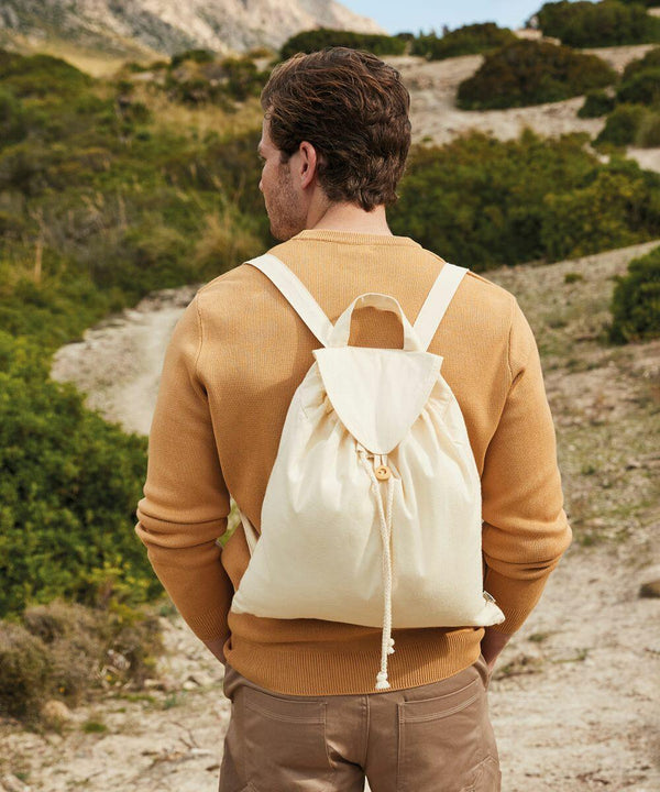 A person wearing a beige sweater and tan pants stands outdoors on a dirt path with green bushes, carrying a beige Westford Mill WM185 drawstring backpack.