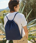 Person standing outdoors wearing a white shirt and carrying a dark blue WM210 drawstring backpack from Westford Mill, with large green plants and a fence in the background.