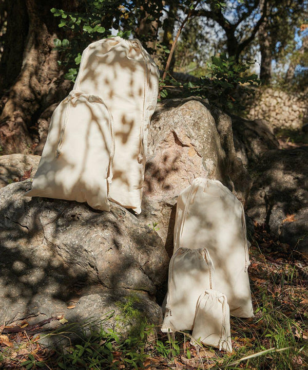 Several Westford Mill WM216 drawstring bags of different sizes are placed on and around large rocks in a wooded outdoor setting.