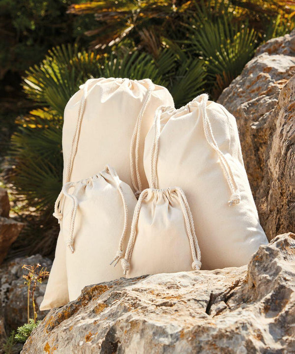 Several Westford Mill WM266 drawstring fabric bags of varying sizes are placed on rocky ground with green plants in the background.