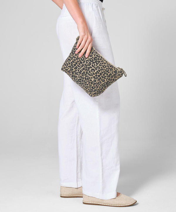 Against a plain, light background, a person in white linen trousers and beige slip-ons holds the Westford Mill Leopard print canvas accessory bag.