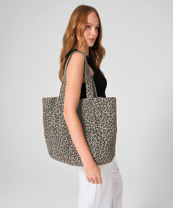 A woman with long red hair wears a sleeveless black top and white trousers, holding the Westford Mill Leopard Print Canvas Shopper over her shoulder, standing against a plain light grey background.