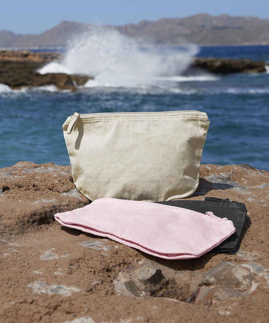 The Westford Mill garment-dyed accessory bag stands upright on rocks near the sea, with pink and black folded fabric pieces in front. Waves crash against the shore and distant mountains are visible in the background.