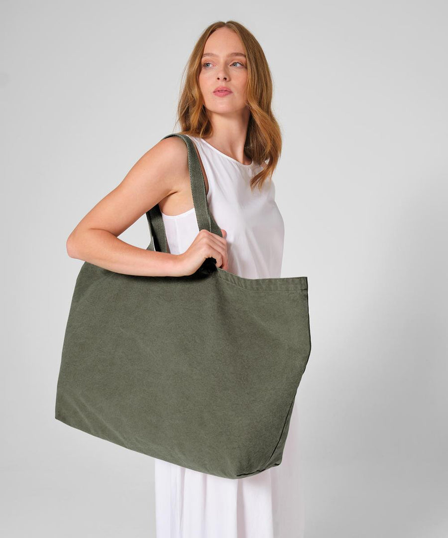 A woman with long, light brown hair in a sleeveless white dress holds the Westford Mill garment-dyed oversized tote bag in olive green over her shoulder against a plain light grey background.