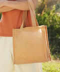 A person in an orange shirt and beige pants is holding a tan, rectangular tote bag with wide handles, Westford Mill WM406, standing outdoors.