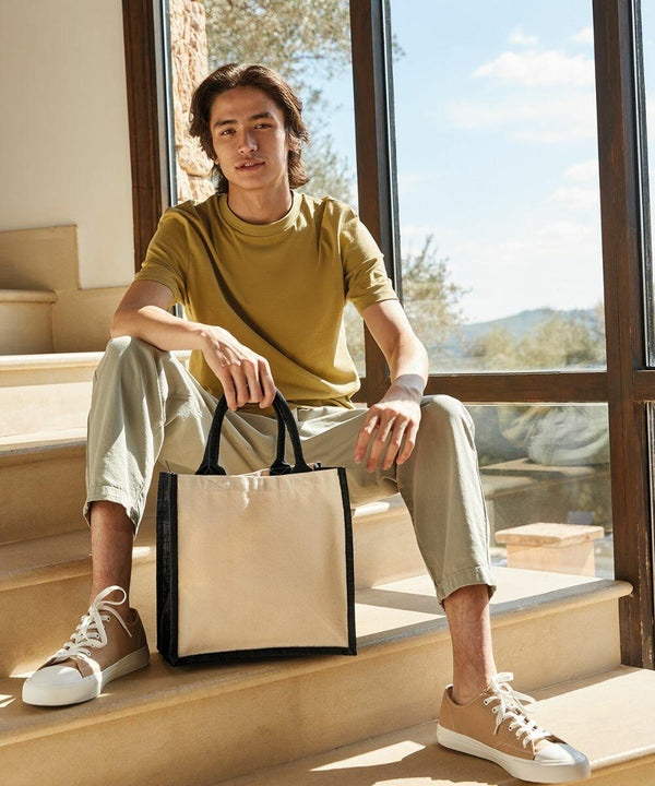 A person sits on indoor stairs wearing a yellow T-shirt, light-colored pants, and beige sneakers, holding a Westford Mill WM421 tote bag with black handles. Sunlight filters through the large window behind them.