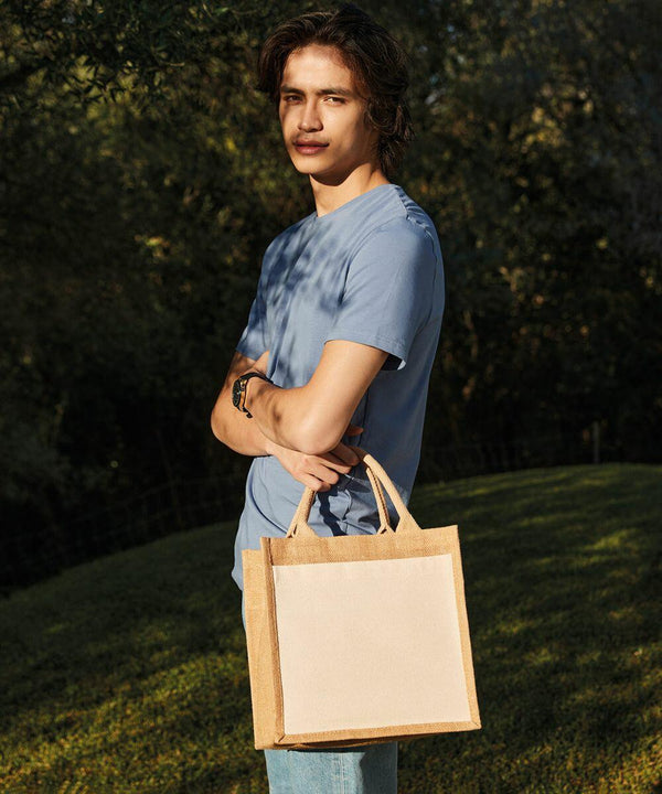 Person standing outdoors holding a beige and brown Westford Mill WM426 reusable tote bag with both hands, wearing a light blue shirt and blue jeans. Greenery is visible in the background.