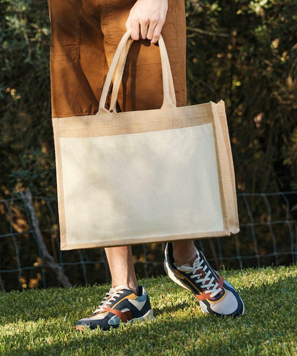 Person carrying a beige Westford Mill WM427 tote bag with tan handles, wearing brown pants and multi-colored sneakers, walking on a grassy area with a wire fence in the background.