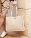 Person holding a large beige Westford Mill WM447 tote bag by its handles, standing next to a light-colored wall.